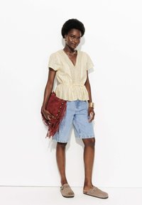 Blouse peplum en beige clair avec des détails en dentelle, assortie à un short en denim bleu Bermuda. Accessoirisée avec un sac à franges rouge et des sabots beiges.