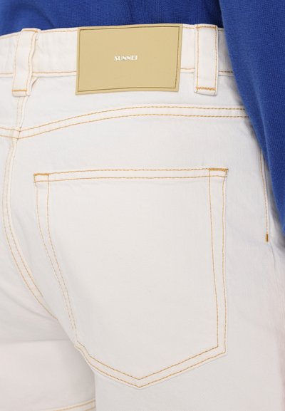 Pantalones cortos de mezclilla blanca con costuras naranjas. Cuenta con un bolsillo cosido y una etiqueta con "SUNNEI" sobre un fondo beige.