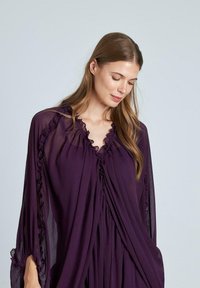Fioletowa sukienka z szyfonu z falbankami przy dekolcie i rękawach, z drapowanym materiałem nadającym płynny kształt. Gładka tekstura.