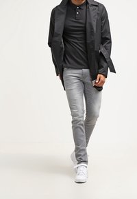 Zwarte lichtgewicht jas over een zwart polo-shirt, gecombineerd met slim-fit grijze jeans en witte sneakers, wat een casual look benadrukt.