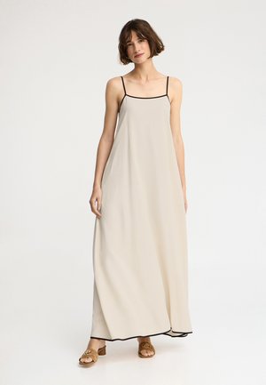 Kobieta z krótkimi brązowymi włosami, ubrana w luźną beżową suknię maxi z czarną lamówką i brązowe sandały na obcasie, stojąca na jednolitym białym tle.