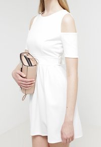 Vestido blanco de manga corta con hombros al aire y tejido texturizado, combinado con un bolso de mano beige con cierre y detalles en negro.