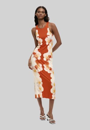 Donna in piedi con un abito midi arancione senza maniche, aderente, con grandi stampe floreali bianche e gialle e sandali bianchi con tacco.