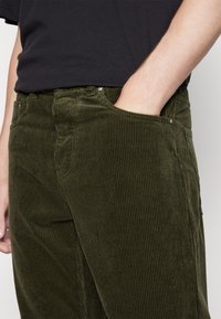 Groene flanellen broek met opvallende verticale ribbels, een rechte snit en zijzakken; details zijn onder meer zilverkleurige hardware en een gestructureerde afwerking.