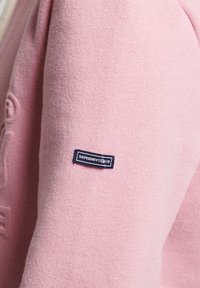 Jersey rosa claro con una textura suave. Presenta una etiqueta azul marino con "Superdry" cosido en la manga. Diseñado para la comodidad y el uso casual.