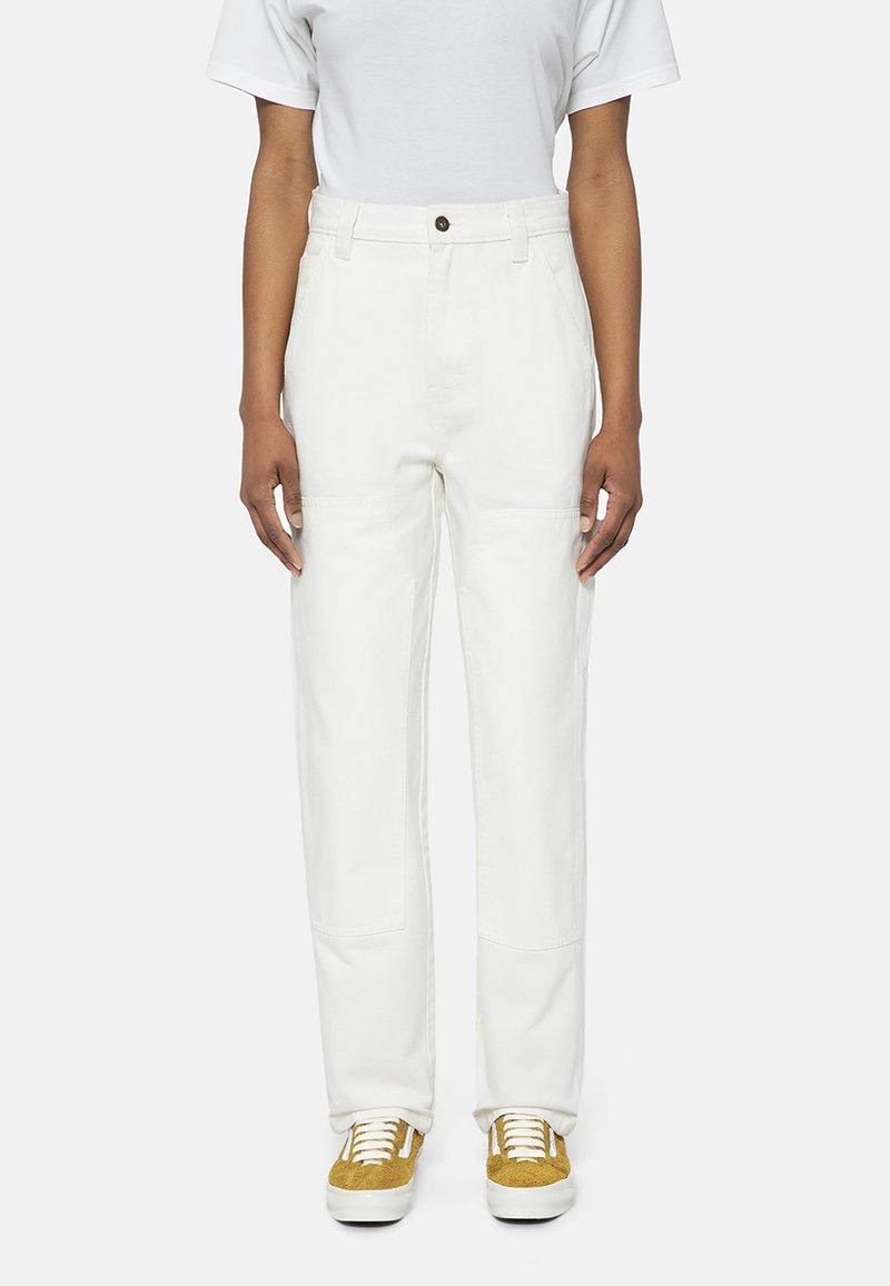 Personne portant un pantalon blanc taille haute, une chemise blanche à manches courtes et des baskets jaunes et blanches, debout devant un fond uni.