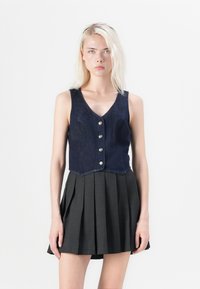 Denim cropped vest met een V-hals, voorzien van vijf zilveren knopen. Gecombineerd met een geplooide, donkergrijze rok, wat een gelaagd uiterlijk creëert.