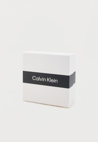 Weißer rechteckiger Karton mit einem schwarzen Band, das den Text "Calvin Klein" in weißer Schrift zeigt. Glatte Textur, minimalistisches Design.