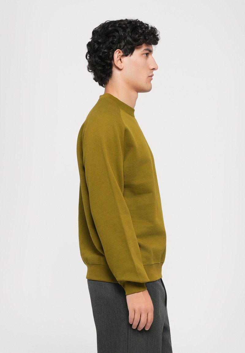 Jeune homme aux cheveux bouclés et foncés portant un sweat-shirt à manches longues vert olive et un pantalon gris, se tenant de profil devant un fond uni.