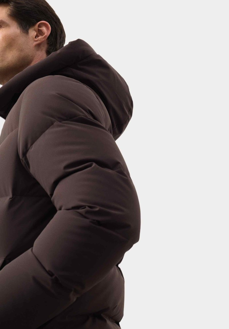 Brązowa kurtka puchowa z kapturem, z pikowanymi sekcjami i gładką teksturą, pokazana z boku, aby podkreślić jej design i kształt.