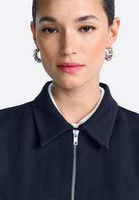 Sötétkék dzseki strukturált gallérral és ezüst cipzárral, kör alakú ezüst fülbevalókkal párosítva. Sima textúra világos háttér előtt.