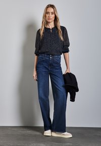 Schwarze gemusterte Bluse mit Knopfleiste, hochtaillierte blaue Jeans und weiße Sneakers. Das Outfit wird mit einer dunklen Jacke in der Hand abgerundet.