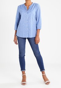 Chemise boutonnière bleu clair avec des motifs abstraits blancs, manches longues et décolleté en V, associée à un jean skinny bleu foncé et des sandales claires.