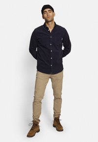 Marineblaues Cordhemd mit langen Ärmeln, hellbraune Hosen und braune Schnürstiefel. Verfügt über eine Brusttasche und ein minimalistisches Design.