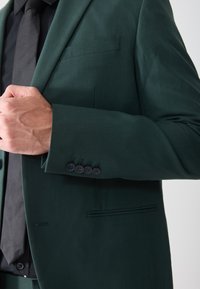 Dunkelgrünes Sakko aus glattem Stoff mit einer einzelnen Brusttasche und einem Detail mit vier Knöpfen an den Manschetten. Eine schwarze Krawatte ist teilweise sichtbar.