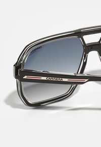 Ochelari de soare din plastic negru, cu lentile gri în degrade. Prezintă un cadru unghiular distinctiv și un logo metalic cu accente roșii pe tempă.
