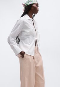 Γυναίκα με μακριά πλεξούδα, που φοράει λευκό πουκάμισο με δέσιμο μπροστά, μπεζ παντελόνι φαρδύ και παστέλ μαντήλι με σχέδια στο κεφάλι, στέκεται με τα χέρια στις τσέπες.