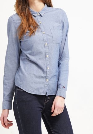 Woman wearing a light blue button-up shirt with a chest pocket and dark blue jeans, standing against a white background.
