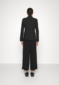 Woman with hair in bun wearing black blazer, wide-leg pants, and black platform shoes standing facing a white wall.