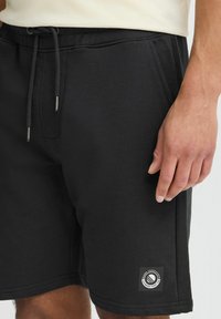 Shorts noirs avec une taille à cordon, dotés d'une texture douce et d'une étiquette logo sur le côté gauche. Design épuré et coupe décontractée.