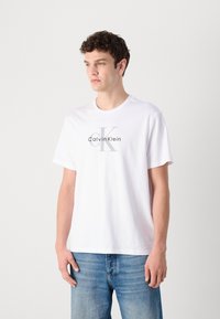 Bijela pamučna majica s velikim sivim logotipom Calvin Klein i 'CK' u podebljanom fontu. Jednostavna okrugla izreza i klasične kratke rukave.