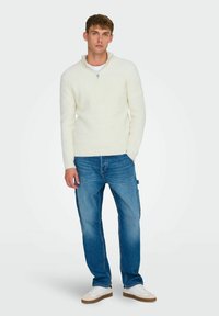 Krämfärgad ribbad tröja med dragkedja i kragen, tillsammans med ljusblå jeans och vita sneakers. Tyget ser mjukt ut med en texturerad yta.