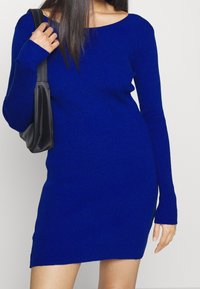 Modrá pletená šaty s žebrovanou texturou, přiléhavý silueta a dlouhými rukávy. Modelka drží černou tašku a pózuje na neutrálním pozadí.