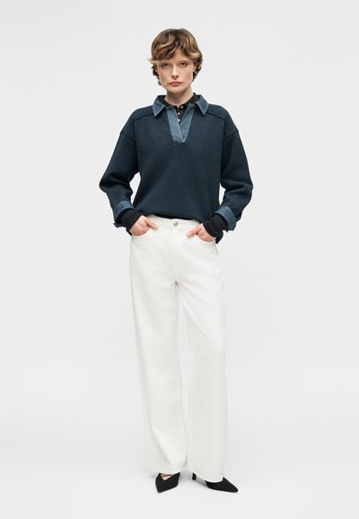 Άτομο που στέκεται με τα χέρια στις τσέπες, φορώντας σκούρο μπλε πουλόβερ με γιακά και μανσέτες από τζιν, λευκό παντελόνι με φαρδιά μπατζάκια και μαύρες μυτερές γόβες.