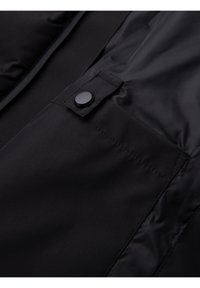 Veste en tissu noir en gros plan montrant une poche avec un rabat sécurisé par un bouton pression noir rond.