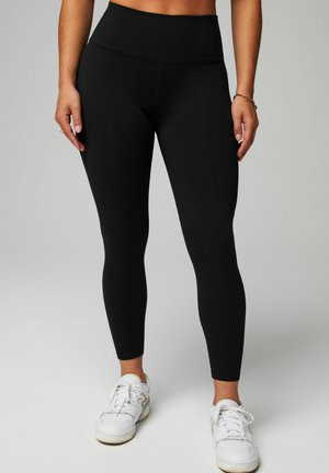 Leggings noirs taille haute en tissu extensible, avec une texture lisse et un design ajusté, associés à des chaussures de sport blanches.