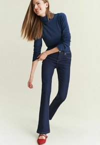 Marinefarbener Rollkragenpullover, kombiniert mit dunkelblauen Schlagjeans. Rote Ballerinas setzen einen Akzent im Outfit. Glatte Texturen und eine figurbetonte Passform sind besonders auffällig.