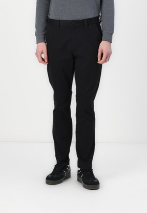 Mand iført sorte bukser, sorte sneakers og en grå langærmet skjorte står mod en hvid baggrund.