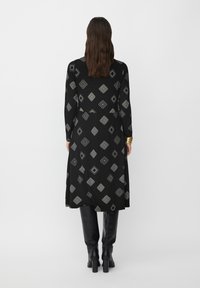 Robe noire à manches longues avec un motif géométrique argenté, coupée au genou, texture douce, associée à des bottes noires montantes.