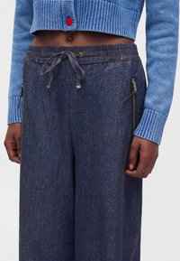 Pantalon large bleu foncé à taille ajustable avec cordon et poches latérales zippées, porté avec un pull court bleu laissant apparaître le ventre.