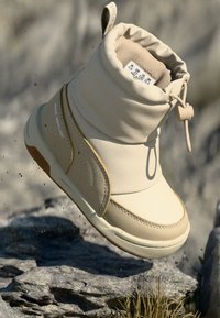 Beige waterdichte boot met een gewatteerde boord, gladde textuur, contrasterende tan zool en elastische sluiting. Bevat een logo nabij de bovenkant.