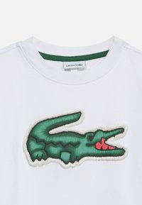Λευκό T-shirt με κεντητό λογότυπο κροκόδειλου σε πράσινο και κόκκινο, κεντραρισμένο κάτω από το λαιμόκοψη, με πράσινη επένδυση στο εσωτερικό του γιακά.