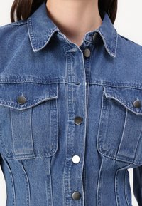 Denim jakna v svetlo modri barvi z gumbi spredaj, koničastim ovratnikom in dvema prsoma žepoma. Ima kovinske gumbe in strukturirane šive.