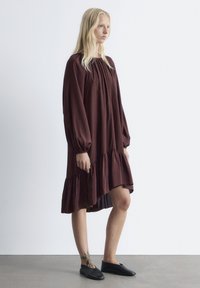 Bordeauxrotes Kleid mit langen, puffigen Ärmeln, gerafftem Ausschnitt und asymmetrischem, geschichteten Saum. Getragen mit schwarzen Slippers.