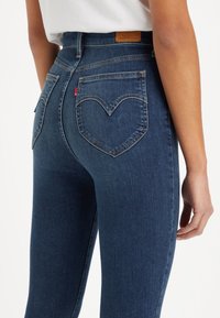 Jeans de mezclilla de cintura alta en azul oscuro con bolsillos traseros en forma de corazón y una etiqueta de cuero marrón en la cintura. Textura suave y diseño ajustado.