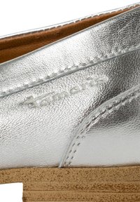 Metallisk silverfärgad sko med texturerad ovandel, som har en sömnadsdesign och en kontrasterande ljusbrun, strukturerad sula. Subtil märkning synlig.