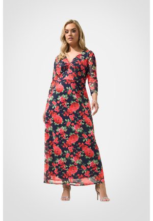 Kvinde iført en lang navyblå kjole med rødt og grønt blomsterprint, trekvartærmer og sølvfarvede højhælede sandaler, stående mod en ensfarvet baggrund.