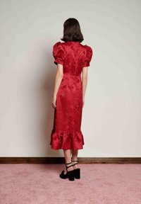 Γυναίκα στέκεται πλάτη, φορώντας κόκκινο σατέν μάκρος φόρεμα με φουσκωτά μανίκια και βολάν στο τελείωμα, συνδυασμένο με μαύρα ψηλοτάκουνα πέδιλα με λουράκια.