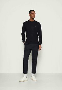 Sweater preto e calças azul-marinho, combinados com ténis brancos. O modelo usa óculos com lentes roseadas, exibindo uma silhueta moderna e ajustada.