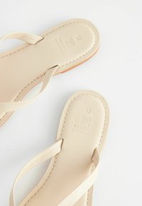 Beige flip-flops med en glat, læderlignende overflade og tynde stropper. Sålen har præget branding og kontrasterende syninger.