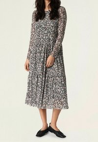 Vestido floral de manga larga con base negra, flores blancas y rosas, un torso ajustado y una falda acampanada. Combinado con zapatos planos negros.