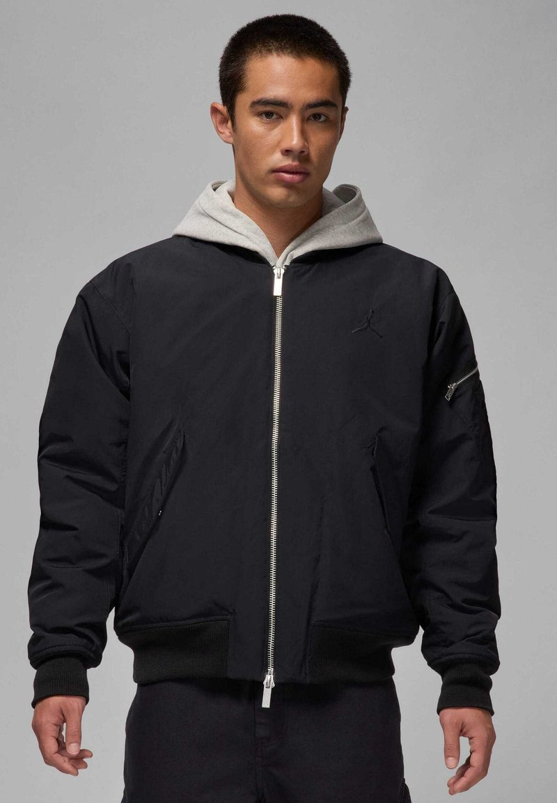 Giubbotto bomber nero con cappuccio grigio, chiusura con zip frontale, polsini e orlo a coste. Presenta una tasca laterale con zip e una discreta personalizzazione sul petto.