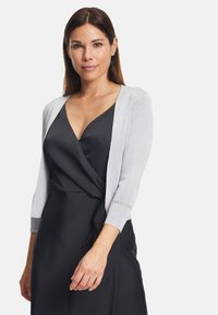 Cardigan grigio chiaro con maniche a tre quarti, con dettagli in lavorazione a maglia aperta sui polsini, indossato su un abito nero. Texture liscia, design aderente.