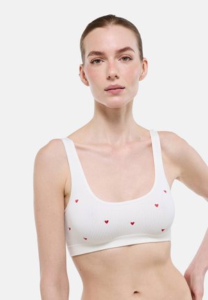 Jeune femme aux cheveux tirés en arrière portant un crop top blanc côtelé avec de petits motifs de cœurs rouges sur un fond uni.