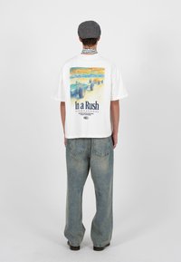 Oseba, oblečena v belo majico z vintage dirkalnimi avtomobili in napisom "In a Rush International" na hrbtni strani, združeno z ohlapnimi kavbojkami in sivim kapo.