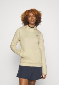 Beige huva sweatshirt i mjukt material med en framficka; har en liten svart bergslogotyp på vänster bröst. Kombinerad med en mörk kjol.